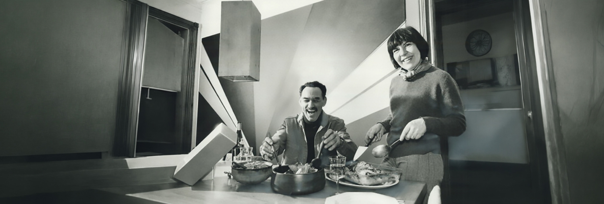 Two people smiling and serving food at a dining table in a modern kitchen, with dishes and drinks placed in front of them.