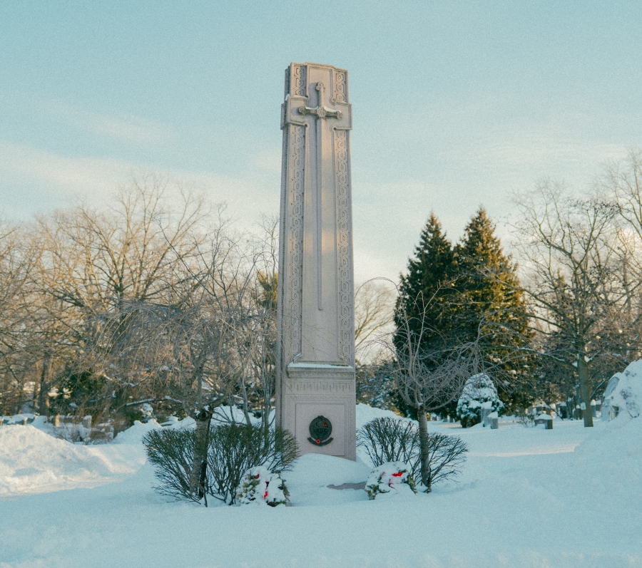 Tall stone war memorial monument standing in a snow‑covered park, surrounded by trees in winter. The monument rises vertically from a landscaped base, with wreaths placed at the foot and bare branches and evergreens in the background.