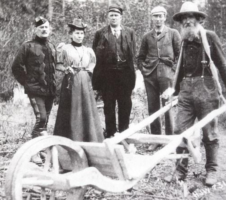 A vintage black‑and‑white photograph showing five people standing outdoors in a wooded area beside a large wooden plow or cart. Four individuals stand in a row wearing period clothing, including long dresses, hats, and suits, while a bearded man in work clothes and suspenders stands to the right holding the wooden equipment. The ground is covered with logs and brush, suggesting a rural or pioneering setting.