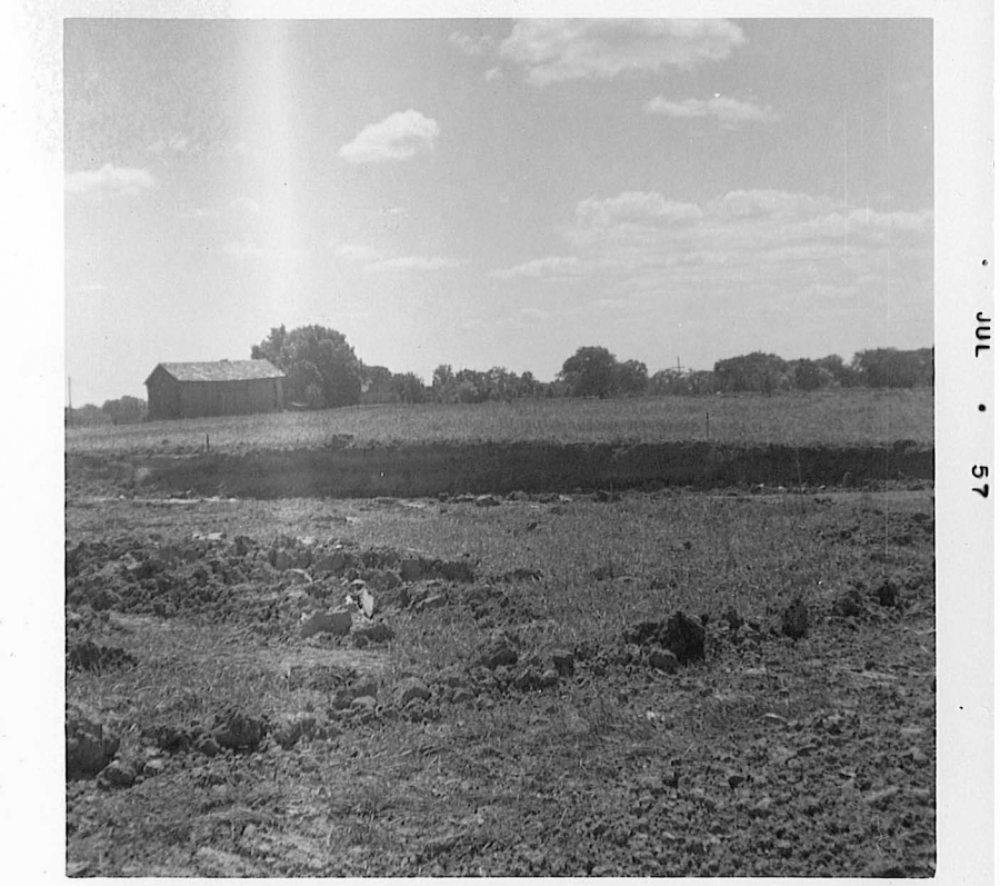 A black‑and‑white photograph of a rural landscape with an open field in the foreground. The ground appears rough and uneven, as if recently plowed. In the middle distance is a flat, grassy area bordered by a line of trees. A small barn or shed sits to the left side of the image. The sky above is partly cloudy. A date mark reading “JUL 57” appears along the right border of the photo.