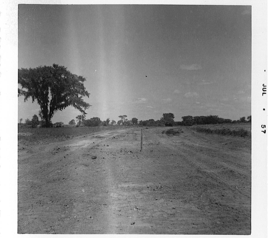 A black‑and‑white photograph of an open, barren dirt field with rough, uneven ground in the foreground. A single wooden stake stands upright near the center of the image, possibly marking a boundary or construction point. Trees and low vegetation line the horizon, with one large tree positioned on the left side. The sky above is partly cloudy. A date mark reading “JUL 57” appears along the right border of the photo.