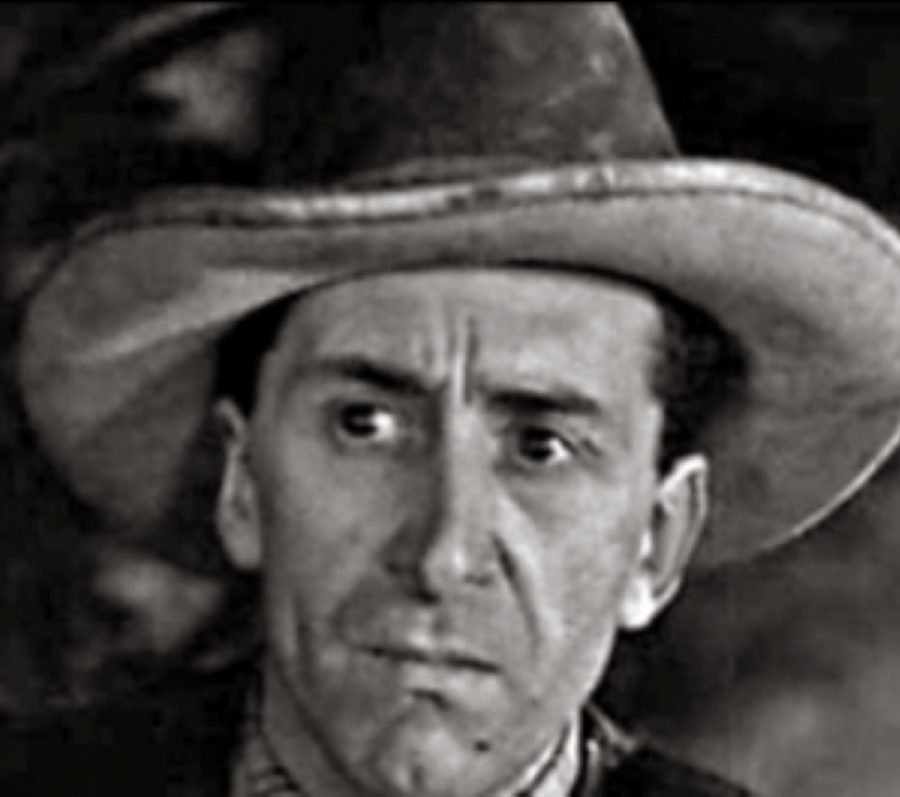 A black‑and‑white close‑up portrait of a person wearing a wide‑brimmed hat, shown from the shoulders up. The background is dark and indistinct, placing emphasis on the hat and upper silhouette.