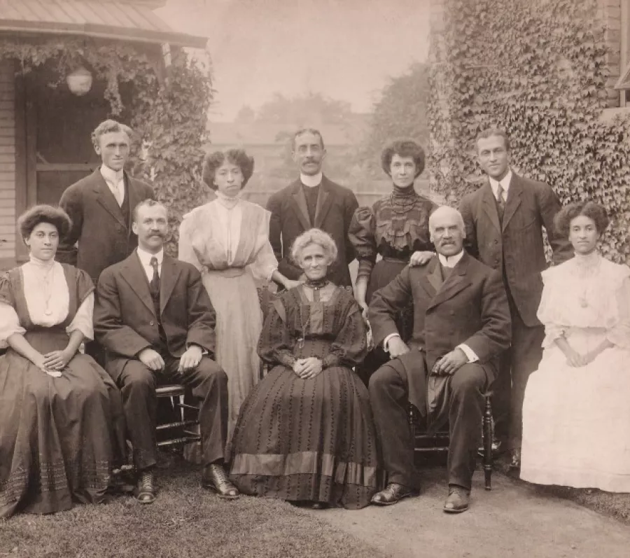 Sepia-toned outdoor group portrait of an extended family posed in front of a house covered in climbing ivy. The group includes adults and children arranged in seated and standing rows. Women wear long dresses with high collars and decorative detailing, while men wear dark suits with ties. A central older woman sits in front, flanked by seated men, with younger and older family members standing behind. The setting and formal attire convey a posed family gathering from the early 20th century.