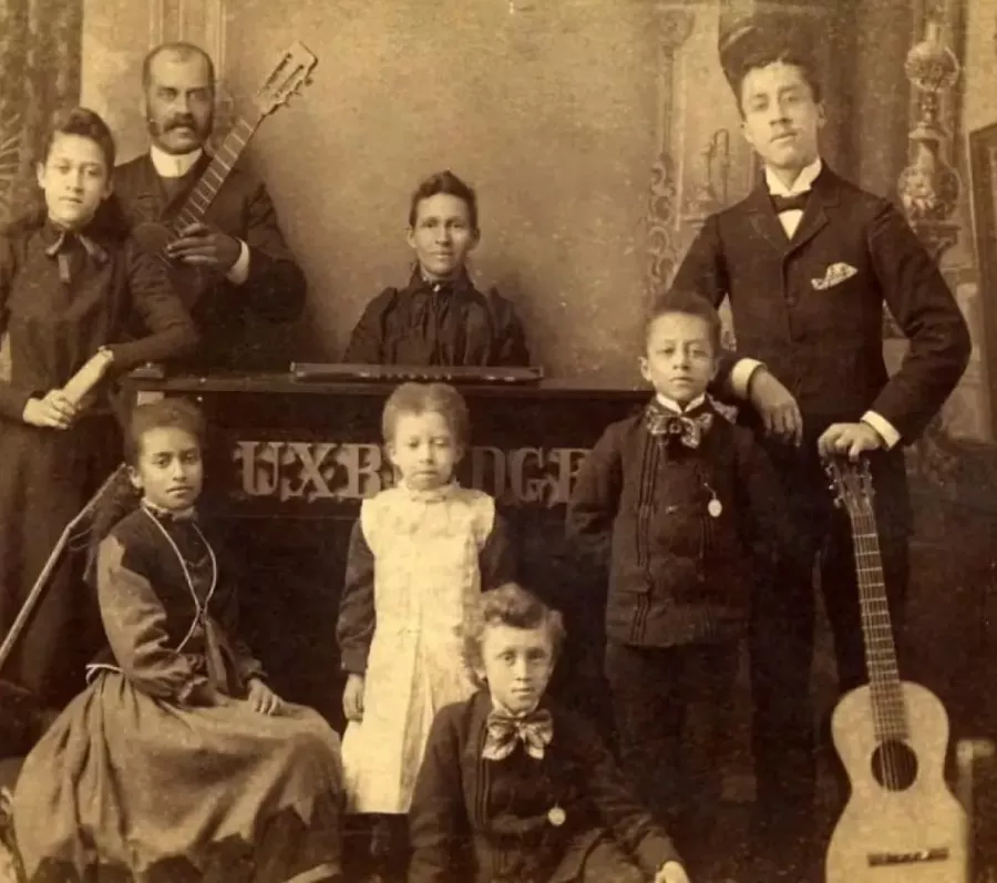 Sepia-toned studio portrait of a large family group arranged around musical instruments and furniture. Adults and children are dressed in formal late-19th- or early-20th-century clothing, including suits, long dresses, and aprons. Several members hold guitars, and a keyboard instrument sits at the center, suggesting a family music group. Ornate painted backdrops and props create a formal indoor setting, conveying a sense of pride, formality, and shared activity.