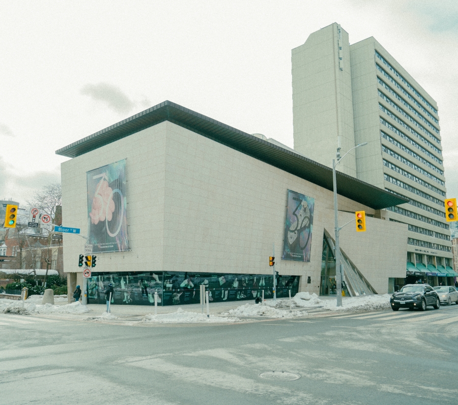 Exterior view of the Bata Shoe Museum, a modern concrete museum building at a city street intersection, with large graphic banners displayed on its walls. Cars and traffic lights are visible at the crossroads, and a tall residential tower rises behind the museum under an overcast sky.