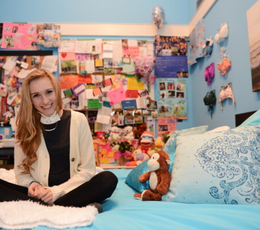 Color photograph of Carley Allison seated cross-legged on a neatly made bed in a bright, cozy bedroom. The room is decorated with light blue walls, patterned pillows, and a small stuffed animal resting on the bed. Behind the bed, a large bulletin board and wall space are densely covered with handwritten notes, cards, photos, and keepsakes. Framed artwork hangs on the walls, and the overall setting feels personal, intimate, and reflective, suggesting a space filled with memories and support.