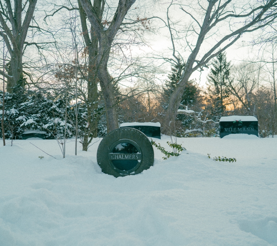 Snow‑covered cemetery scene with a round grave marker in the foreground bearing the name “Chalmers.” The headstone is partially buried in snow, with leafless trees and additional grave markers visible in the background during winter.