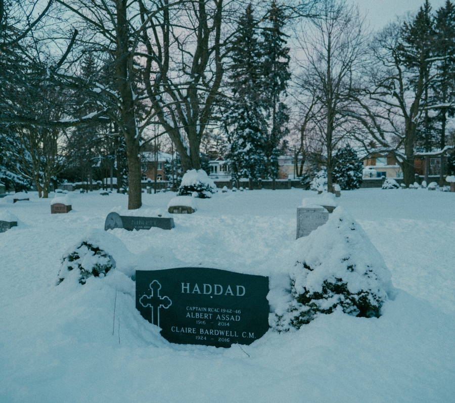 Snow‑covered cemetery scene with several headstones partially buried in snow. A central grave marker in the foreground bears the name “Haddad,” with surrounding trees and residential houses visible in the background during winter.