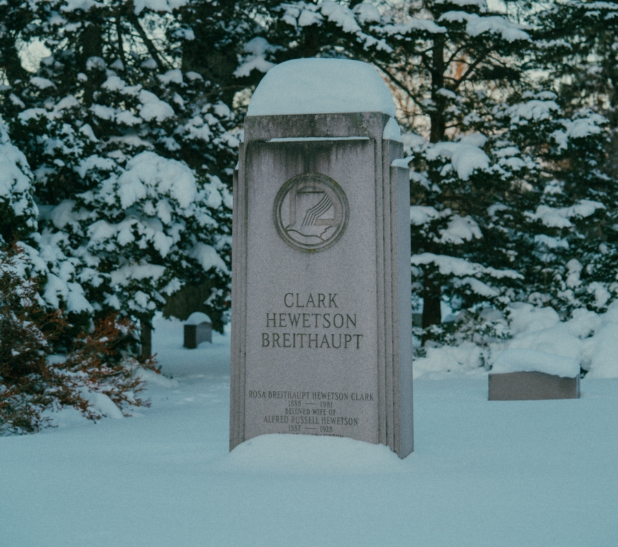 Snow‑covered cemetery landscape with an upright headstone in the foreground bearing the name “Clark Heweston.” The stone is surrounded by trees and additional grave markers, with evergreen and bare branches visible in winter light