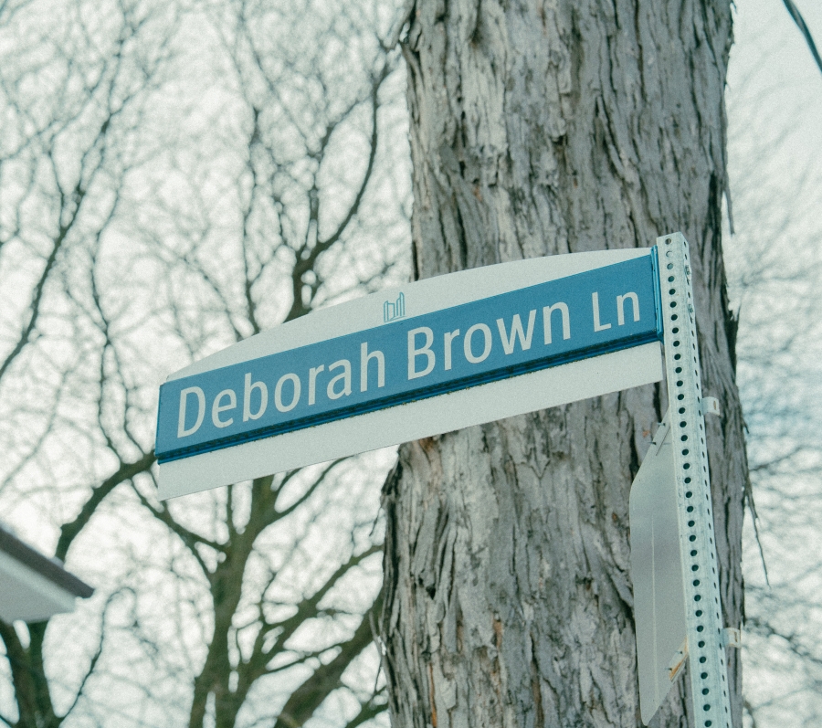 Close‑up of a blue and white street sign reading “Deborah Brown Ln,” mounted on a metal post beside a tree in a residential neighbourhood.