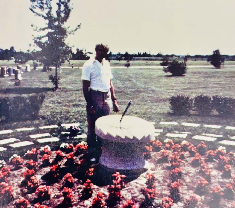 The artist named Sigi Puchta stands beside the sundial. It is a circular stone feature surrounded by red flowers in an open cemetery landscape, with graves, trees, and grassy fields extending into the distance.