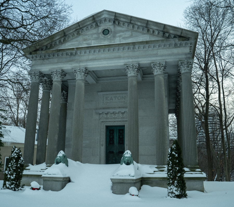 The Eaton Mausoleum, a neoclassical stone structure with tall columns and a triangular pediment, set in a snow‑covered cemetery. Two sculpted lion figures flank the entrance, with bare trees surrounding the mausoleum in winter.