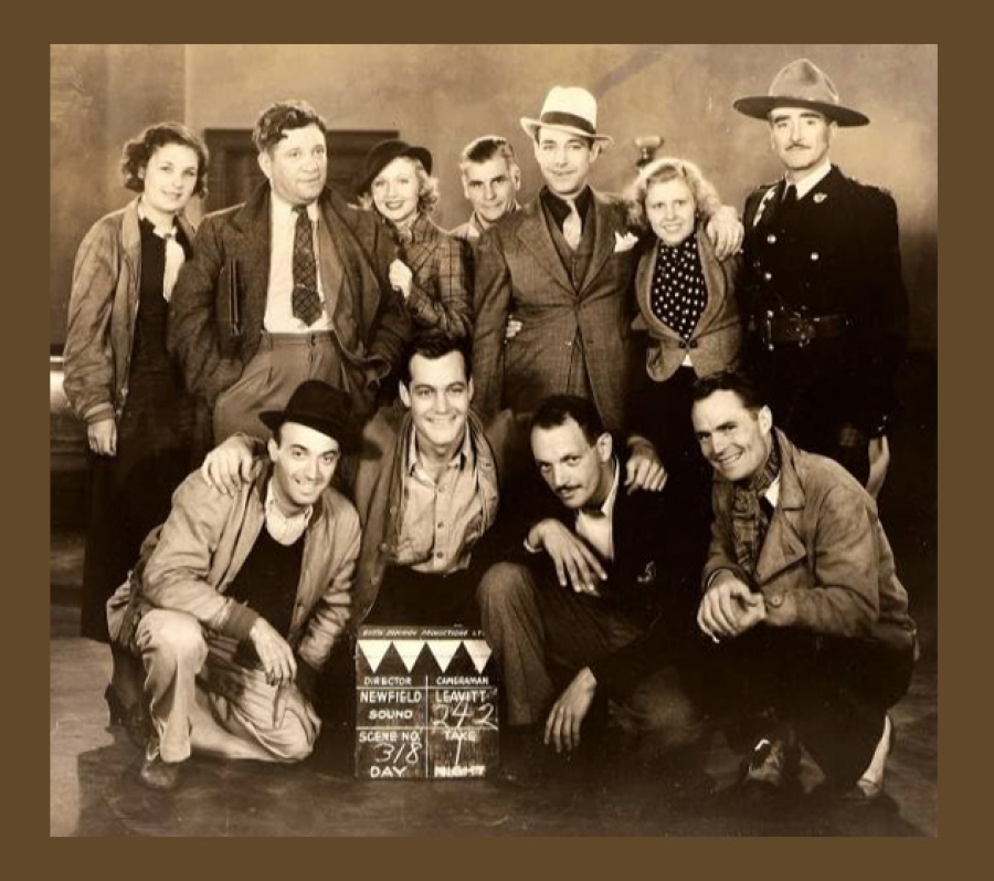 A vintage sepia-toned group photograph featuring several people posed together on what appears to be a film set. Some are standing while others crouch in front. A clapperboard sits near the front row, indicating scene and production details. The group is dressed in period clothing, including suits, hats, work jackets, and a uniformed officer standing to the right. The background suggests an indoor studio environment.