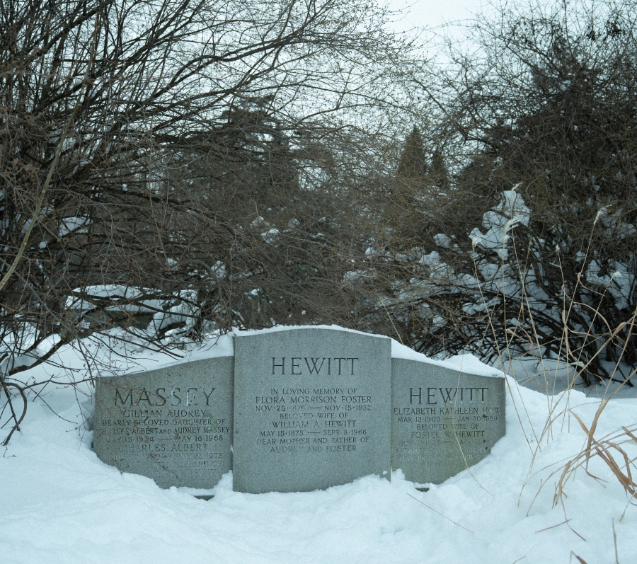 Snow‑covered cemetery scene showing three adjacent grave markers bearing the names Massey and Hewitt. The stones are partially buried in snow and set among dense winter shrubs and leafless trees, creating a quiet, enclosed landscape.
