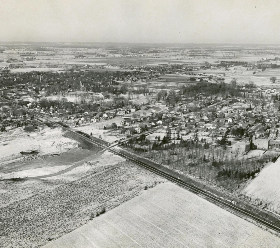 A black and white aerial image of Brampton, taken in 1954. A railroad line can be seen stretching from the middle left edge of the frame to the bottom left corner. In the middle of the frame, there is a concentration of houses and buildings, the landscape dotted with clusters of trees.