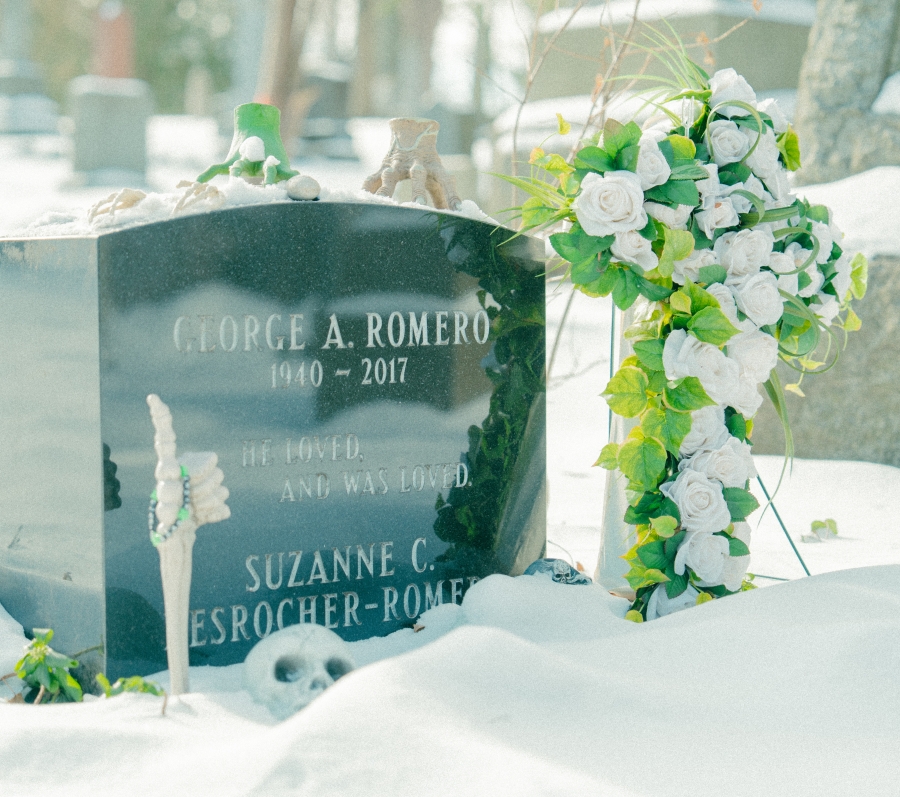 Snow‑covered cemetery scene with a polished black headstone engraved with the name “George A. Romero (1940–2017).” A small skeleton ornament giving a thumbs‑up stands beside the grave, along with a white floral wreath accented with green leaves, with additional headstones visible in the winter background.