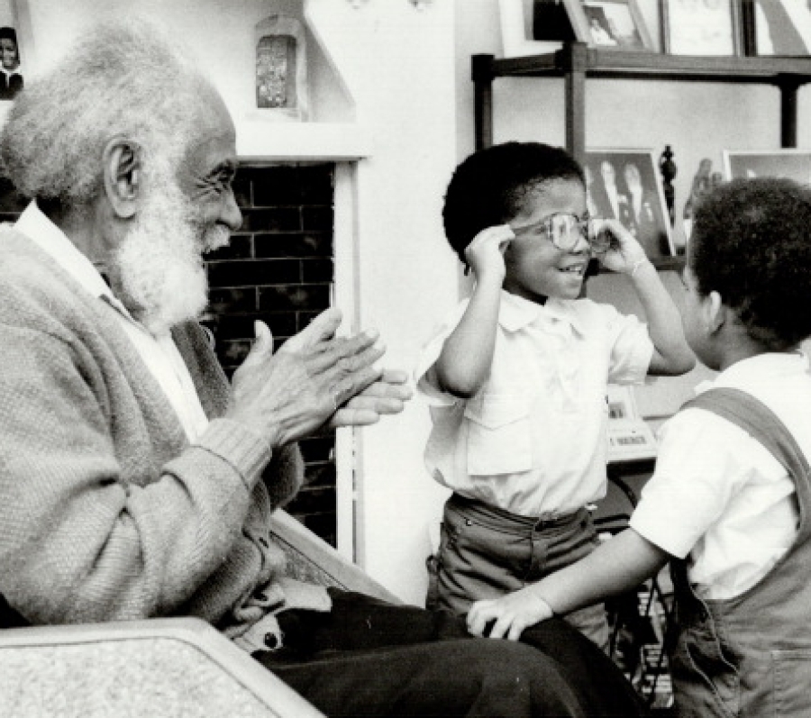 Black-and-white photograph of Harry Gairey seated on a couch in a living room, smiling and clapping as he interacts warmly with two young children standing in front of him. One child raises their hands while the other turns toward Gairey, creating a playful, affectionate moment. Shelves with framed photographs and household items are visible in the background, giving the scene an intimate, home-like feel.
