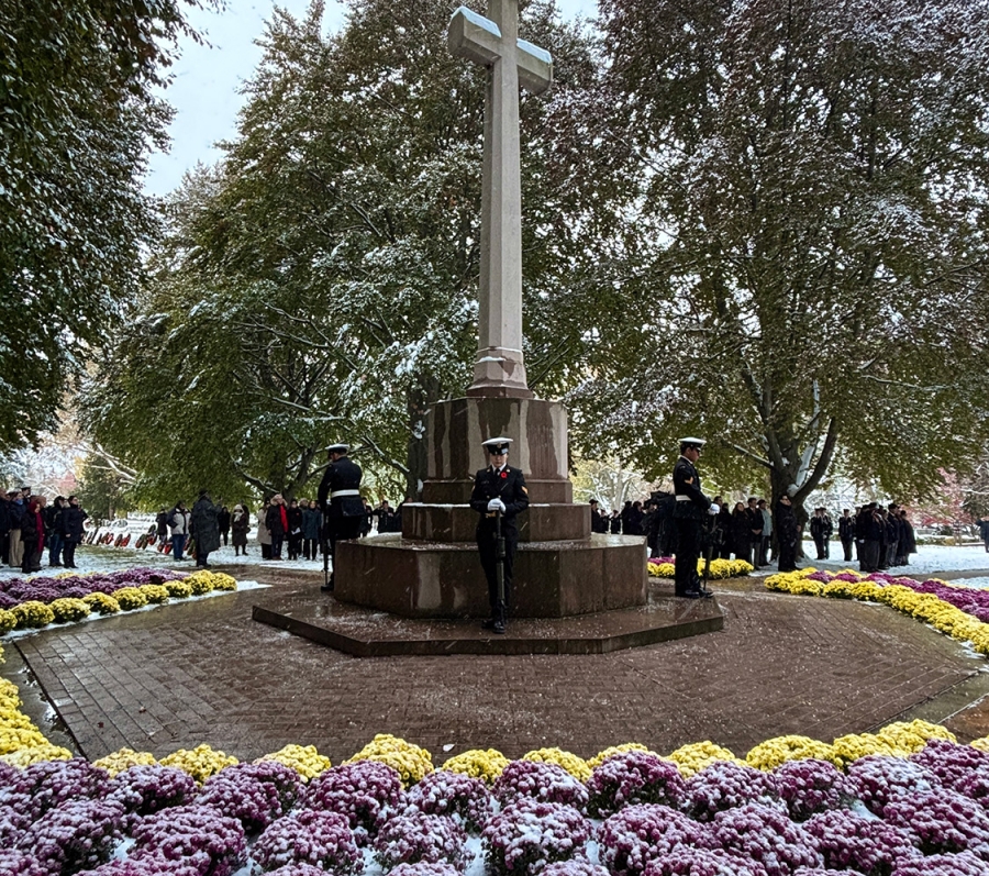 A tall stone cross monument surrounded by snow-dusted flowers, with uniformed guards standing watch and people gathered in the background.