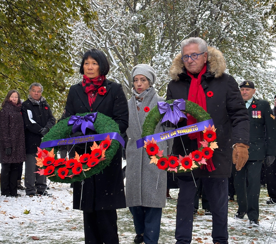 People at an outdoor remembrance ceremony holding wreaths with red poppies and purple ribbons on a snowy day.