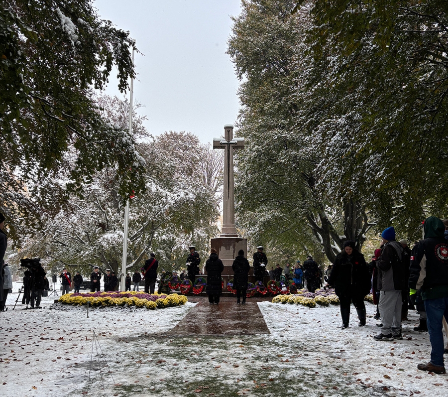 Snowy remembrance ceremony at a war memorial with a large stone cross, wreaths arranged at its base, and people gathered around.