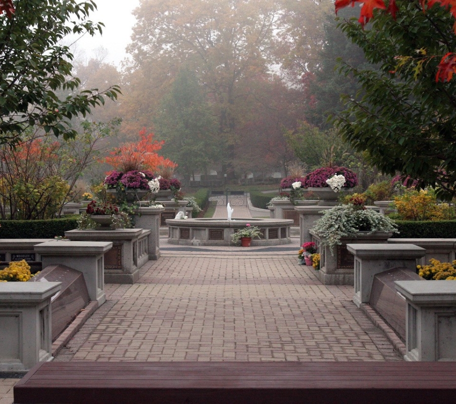 Cemetery garden path with flower-filled planters and central fountain in autumn mist.