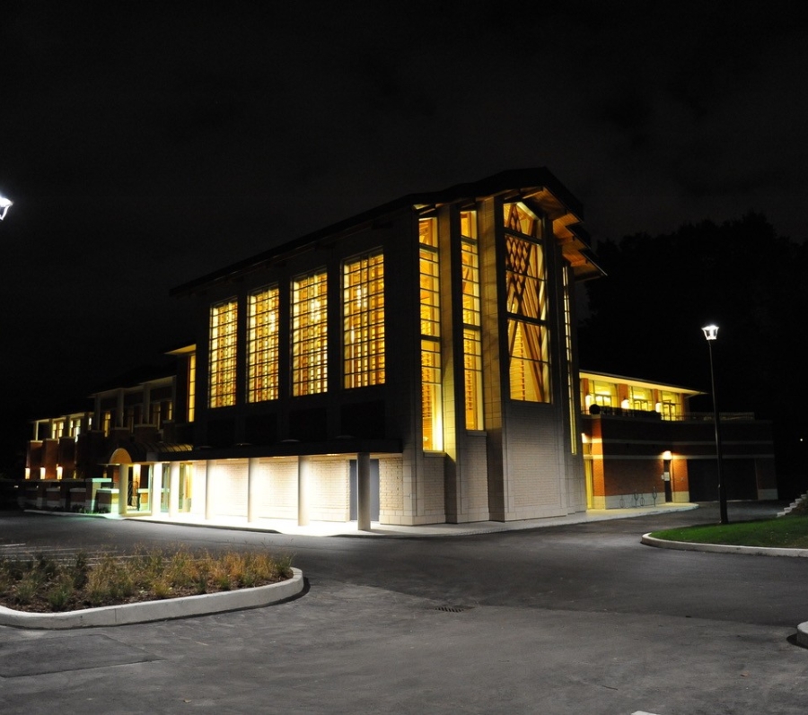 Contemporary building at night with tall lit windows and surrounding pathway and streetlights.
