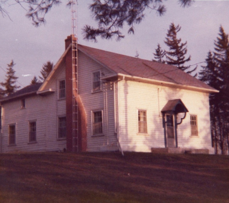 Small white rural building with a sloped roof and chimney, surrounded by trees.