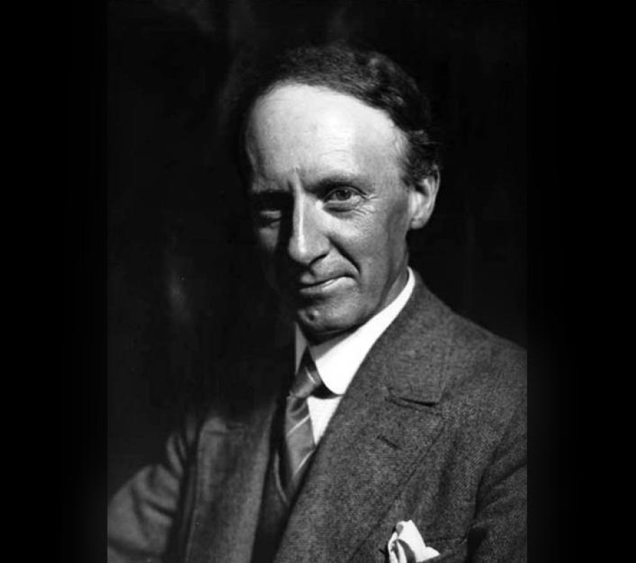 Black‑and‑white vintage portrait of a person wearing a formal suit with a tie and pocket square, seated against a dark studio backdrop.