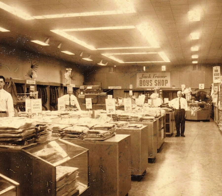 Interior view of a Jack Fraser department store showing long display tables stacked with folded clothing, sales signs hanging above the aisles, and staff standing behind counters under bright ceiling lights.