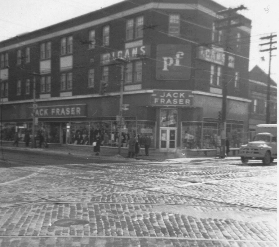 Street‑corner view of the Jack Fraser store at Pape and Danforth, a multi‑storey brick building with large storefront windows, signage along the façade, pedestrians on the sidewalk, and streetcar tracks crossing the intersection in the foreground.