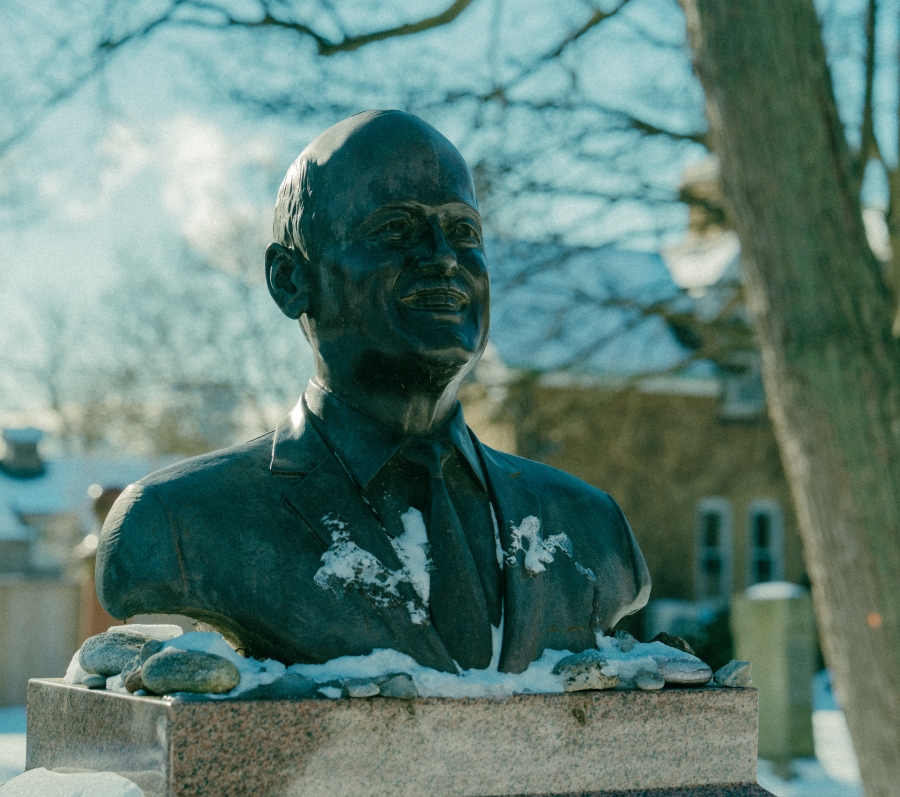 Bronze bust sculpture mounted on a stone pedestal in a snow‑covered cemetery. Snow rests on the shoulders of the sculpture, with bare trees and headstones visible in the winter background.