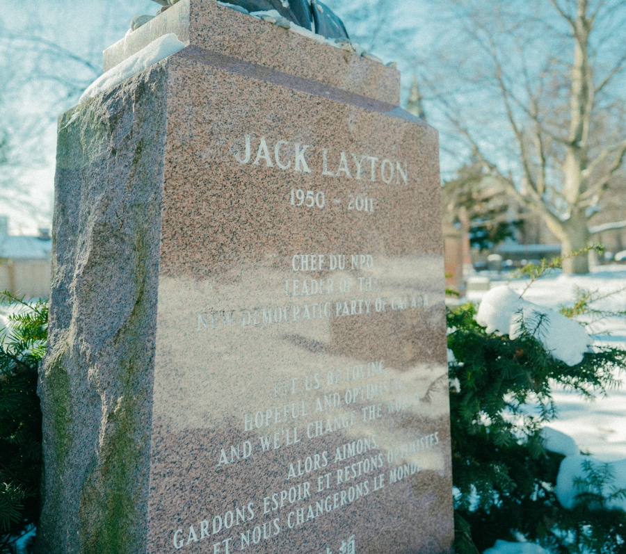 Stone grave marker engraved with the name “Jack Layton (1950–2011)” in a snow‑covered cemetery. Evergreen branches surround the base of the monument, with additional headstones and leafless trees visible in winter light.