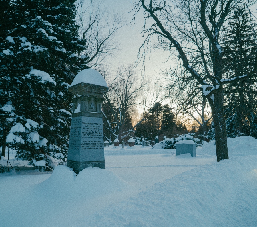 Snow‑covered cemetery scene at dusk featuring a tall stone grave marker in the foreground commemorating James Aikins. The monument is topped with a rounded cap and surrounded by evergreen trees and bare branches, with additional headstones visible along a snow‑covered path in winter light.