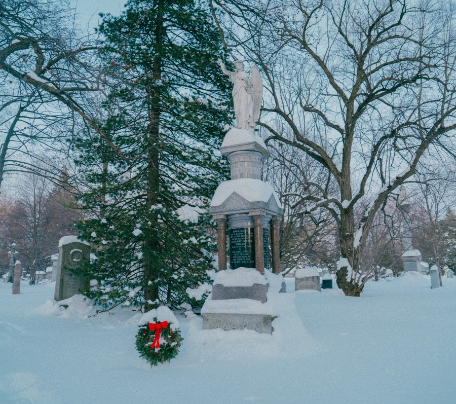 Snow‑covered cemetery scene featuring the Martin family monument, a tall stone memorial topped with a sculpted figure. A wreath with a red bow rests at the base of the monument, surrounded by leafless trees and additional grave markers in a winter landscape.