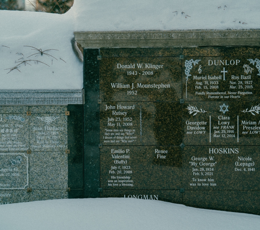 Snow‑covered cemetery scene showing a polished stone memorial wall with multiple engraved name plaques. The central section includes the name “John Rutsey,” with surrounding inscriptions and decorative motifs, partially framed by snow at the base.