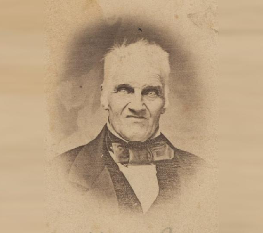 A vintage sepia-toned portrait of a man dressed in formal 19th‑century attire, including a dark coat, waistcoat, white shirt, and large bow tie. The photograph shows age-related wear, such as fading and textured marks, giving it an antique appearance.