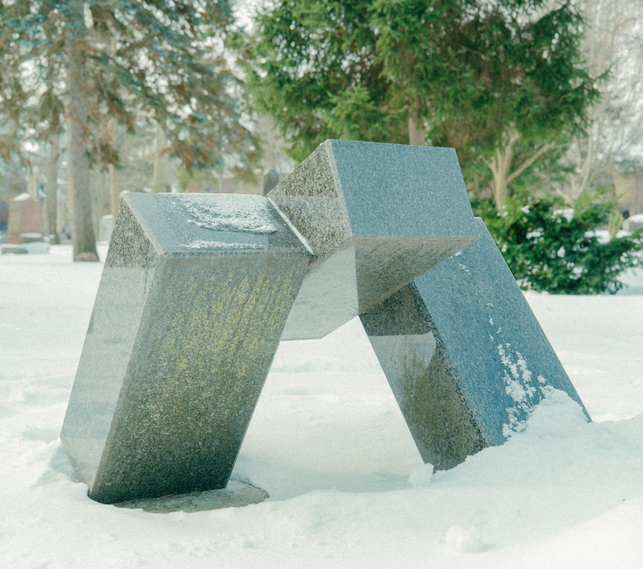 Abstract stone sculpture composed of angled geometric slabs rising from the ground in a snow‑covered cemetery. Trees and surrounding grave markers are visible in the winter landscape.