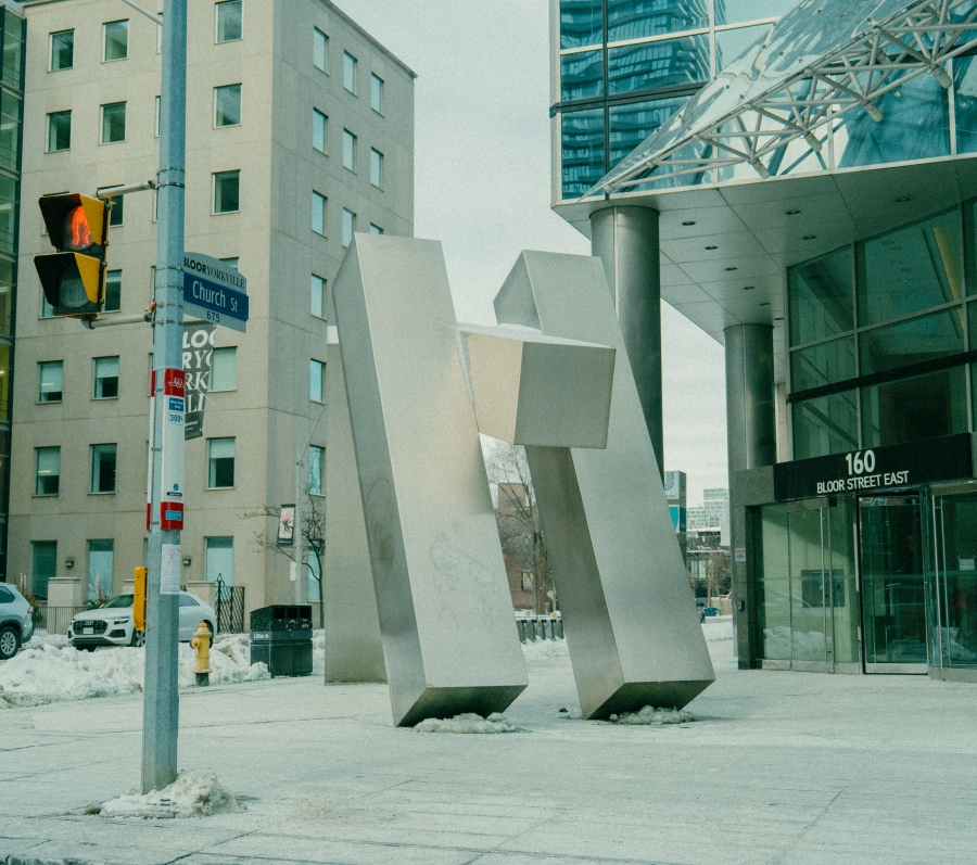 Large abstract metal sculpture made of tall, angular geometric forms installed on a city sidewalk. Modern office buildings, glass façades, and street signage surround the sculpture in an urban downtown setting.