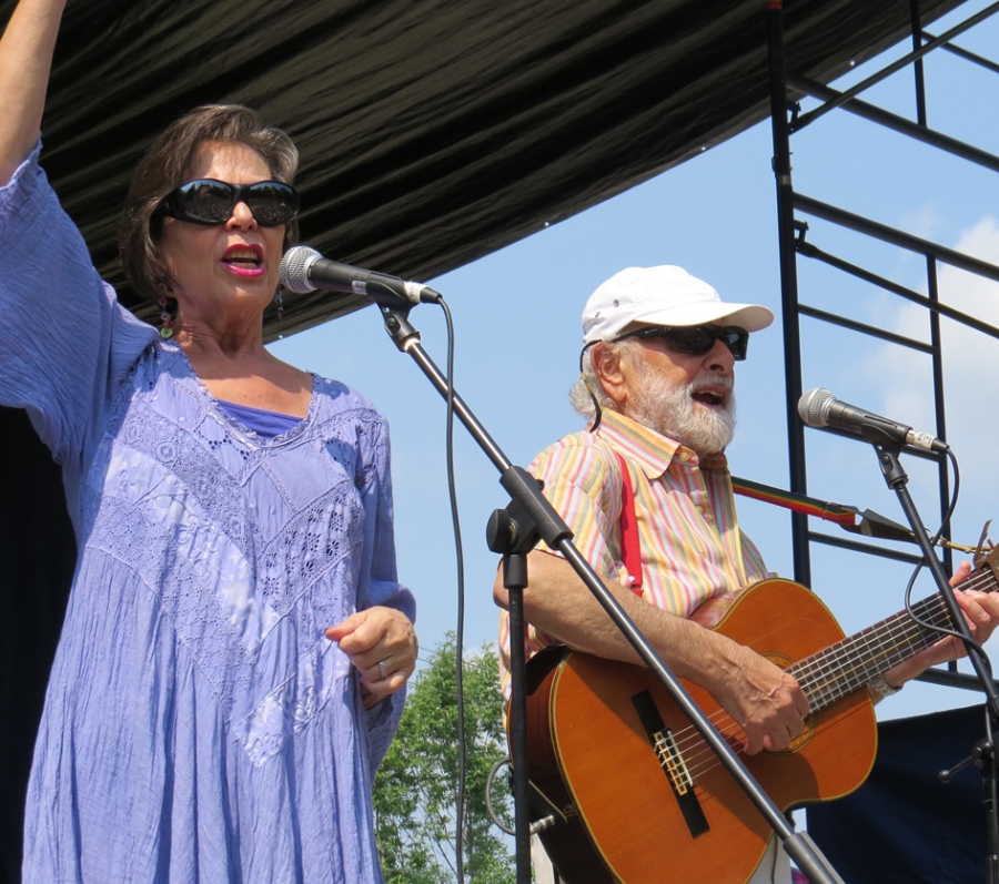 Two performers stand on an outdoor stage; one is singing into a microphone with an arm raised, while the other plays an acoustic guitar beside an additional microphone stand. A canopy and metal stage scaffolding are visible overhead against a sunny sky.