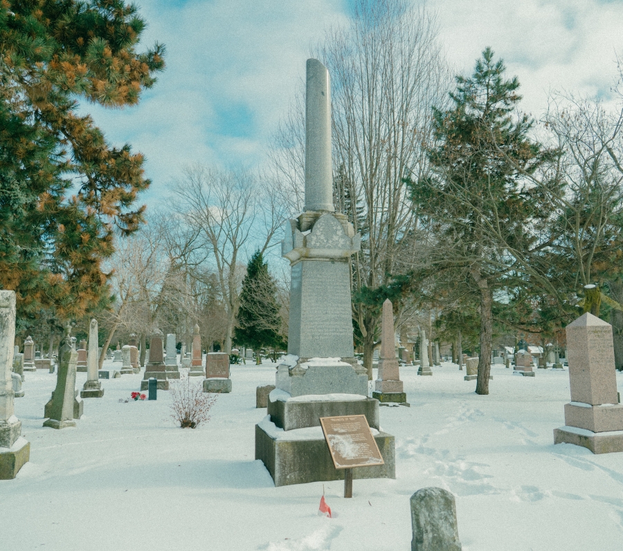 Snow‑covered cemetery landscape featuring the Lount and Matthews monument, a tall stone memorial with a column rising from a stepped base. The monument stands among surrounding headstones and leafless trees in a winter setting, with an interpretive plaque placed in front.