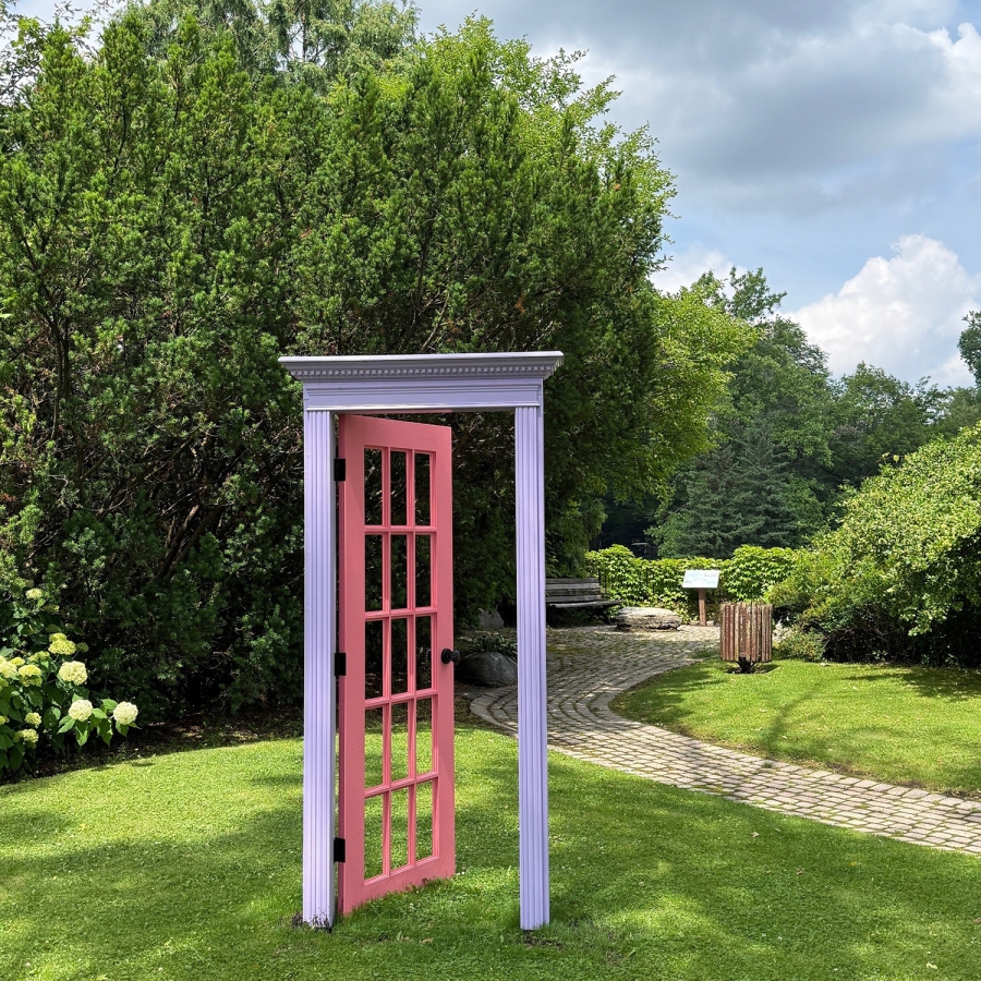 Decorative pink door standing on a lawn beside a garden pathway, surrounded by trees, shrubs, and greenery.