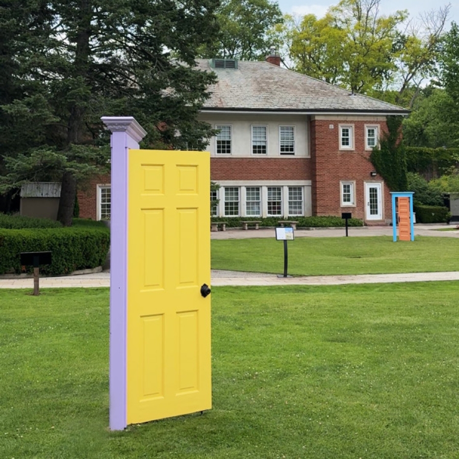 Yellow decorative door standing on a grassy lawn, with a brick building and trees in the background.