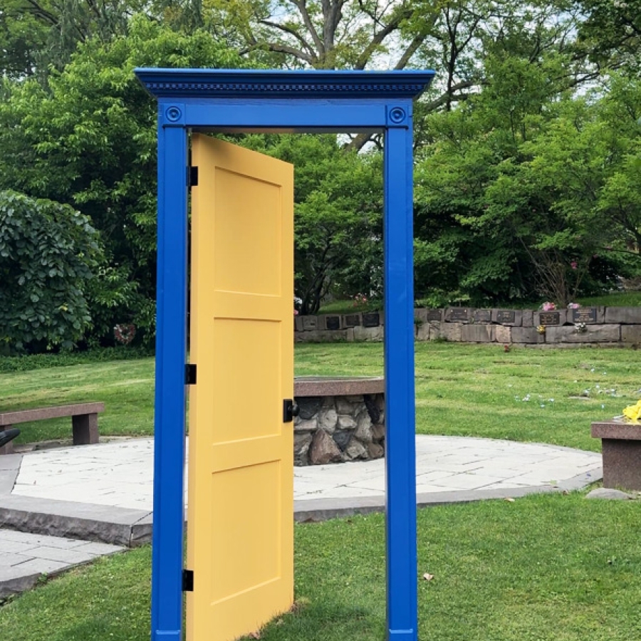 Yellow decorative door within a blue doorframe, standing on a grassy lawn near a circular paved area and surrounded by trees and greenery.