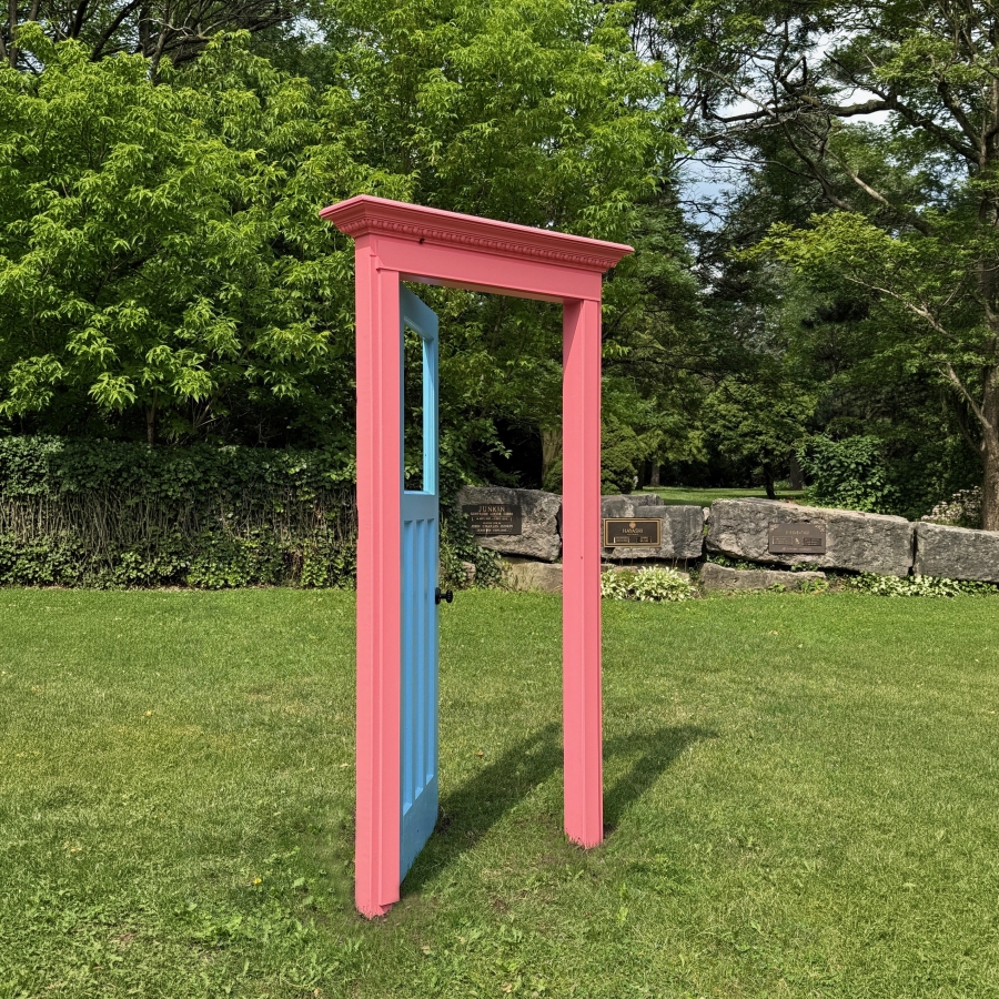 Pink decorative doorframe standing on a grassy lawn, with trees, stone benches, and greenery in the background.