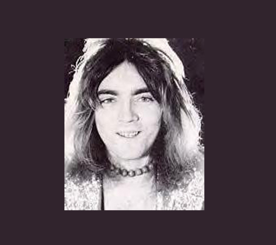 A black‑and‑white portrait of John Rutsey with shoulder‑length hair wearing a beaded necklace and a sequined or textured garment. The lighting creates a soft contrast against the dark background, giving the image a classic studio‑portrait appearance.