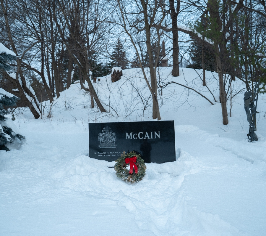 Snow‑covered cemetery scene featuring the McCain family marker, a low black granite headstone set against a wooded hillside. A green memorial wreath with a red bow rests in front of the stone, with trees and snow‑covered ground surrounding the grave in winter.