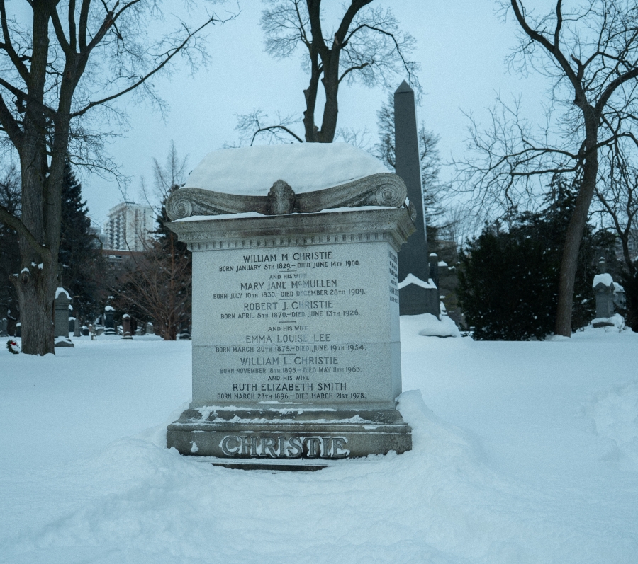 Snow‑covered cemetery scene featuring the McCain family marker, a low black granite headstone set against a wooded hillside. A green memorial wreath with a red bow rests in front of the stone, with trees and snow‑covered ground surrounding the grave in winter.