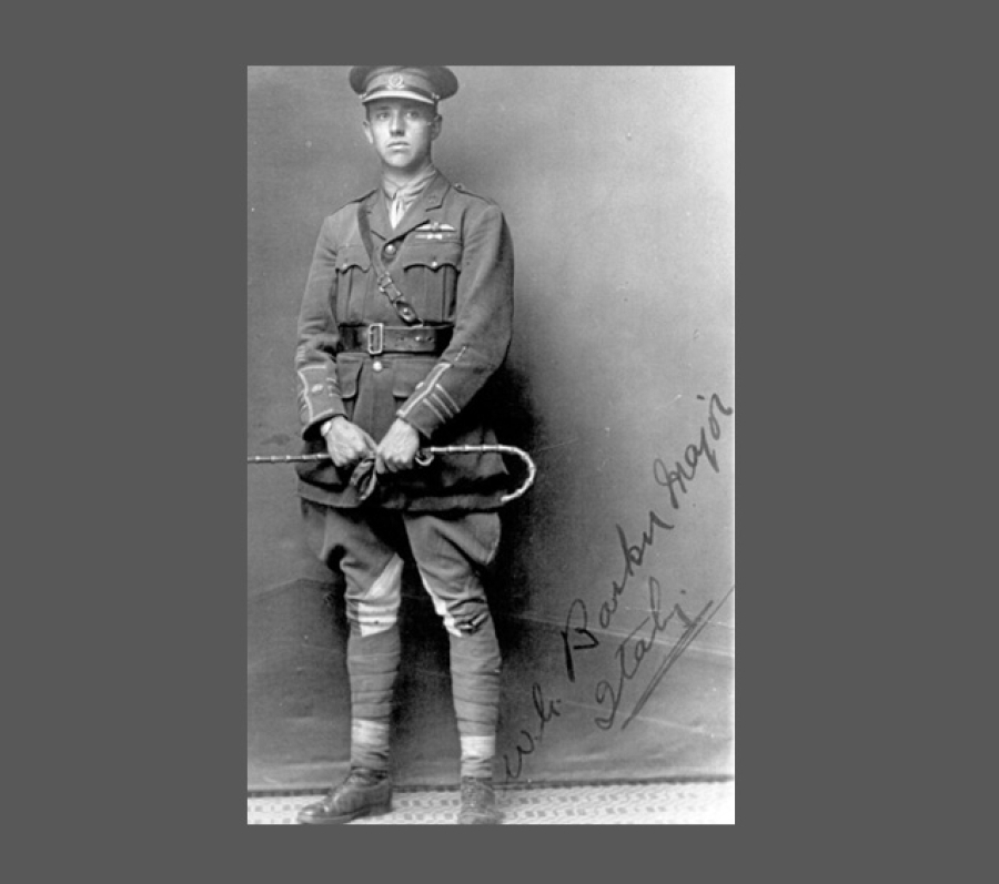 A full‑length black‑and‑white portrait of William George Barker standing in front of a plain studio backdrop. The officer is dressed in early 20th‑century military attire with a belted jacket, medals on the chest, tall boots, and a cap. He holds a riding crop or swagger stick in both hands, positioned in front of him. A handwritten note or signature is visible along the lower right side of the photograph.