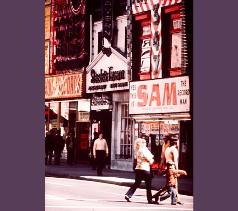 A street scene featuring colorful storefronts, including Sam the Record Man and Steele’s Tavern. Pedestrians walk across the street in the foreground, while others stand near the shop entrances. Bright signage and busy façades create a lively urban atmosphere.
