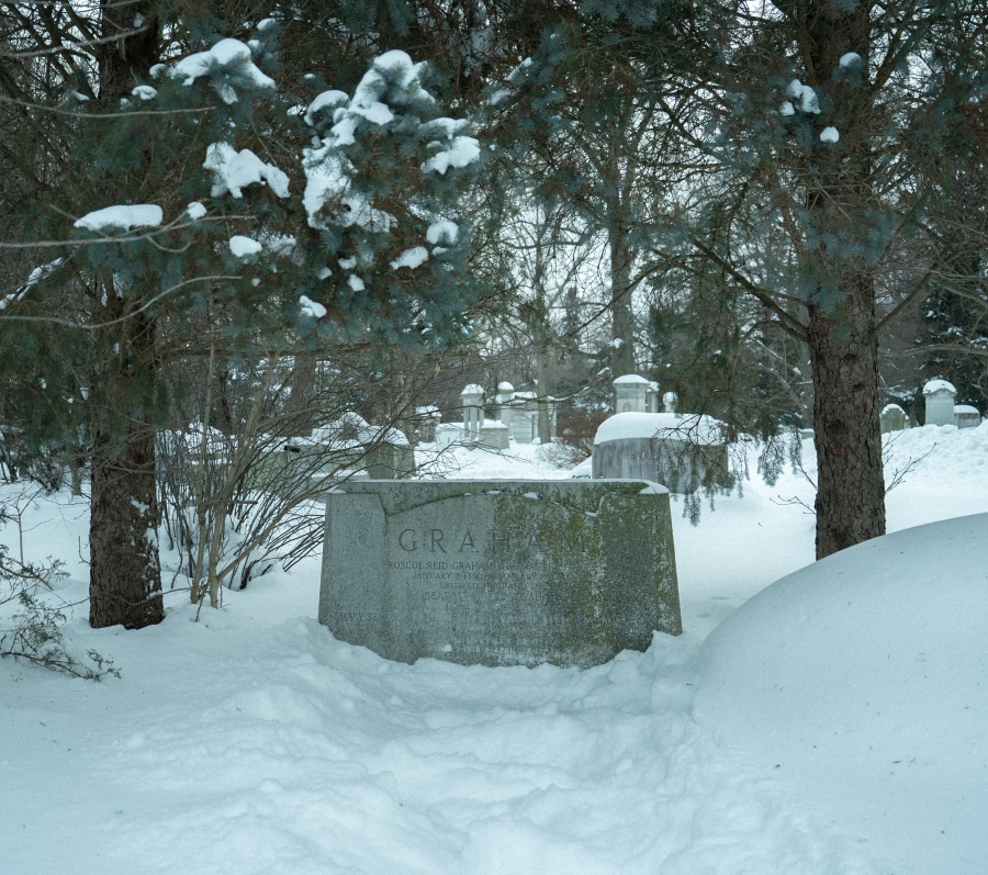 Snow‑covered cemetery landscape with a low, curved stone grave marker partially buried in snow beneath spreading tree branches. Additional headstones and leafless trees are visible in the background, creating a quiet winter scene.